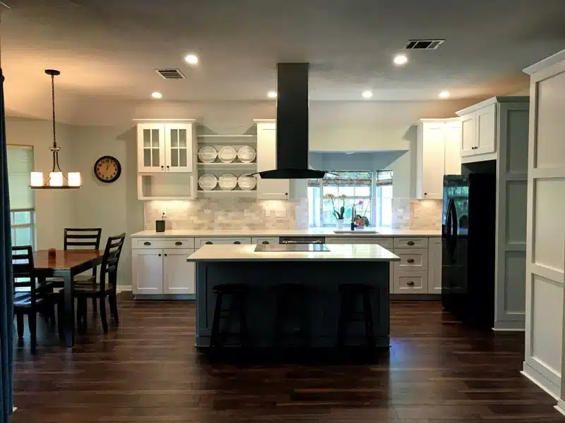 Open kitchen remodel with a gray island, black range hood, white Shaker cabinets, glass-front uppers, and dark wood floors