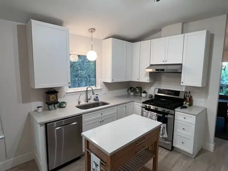 L-shaped kitchen remodel with white Shaker cabinets, light gray countertops, white tile backsplash, and a sink under a window